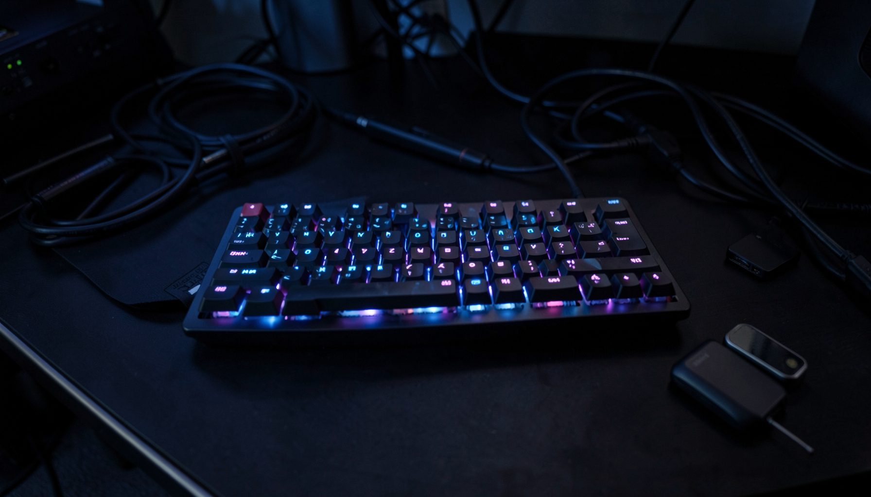 A dark desk surface with a mechanical keyboard glowing in soft blue and purple backlight, surrounded by coiled cables and accessories