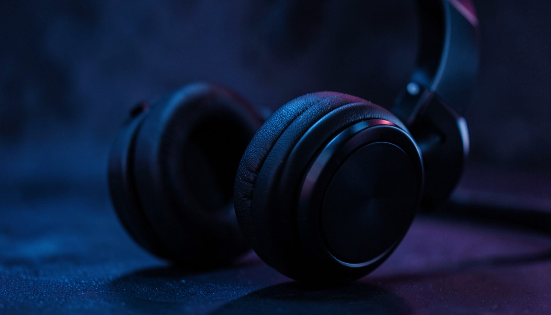 Dark atmospheric close-up of over-ear headphones with soft blue and purple ambient lighting on a moody background