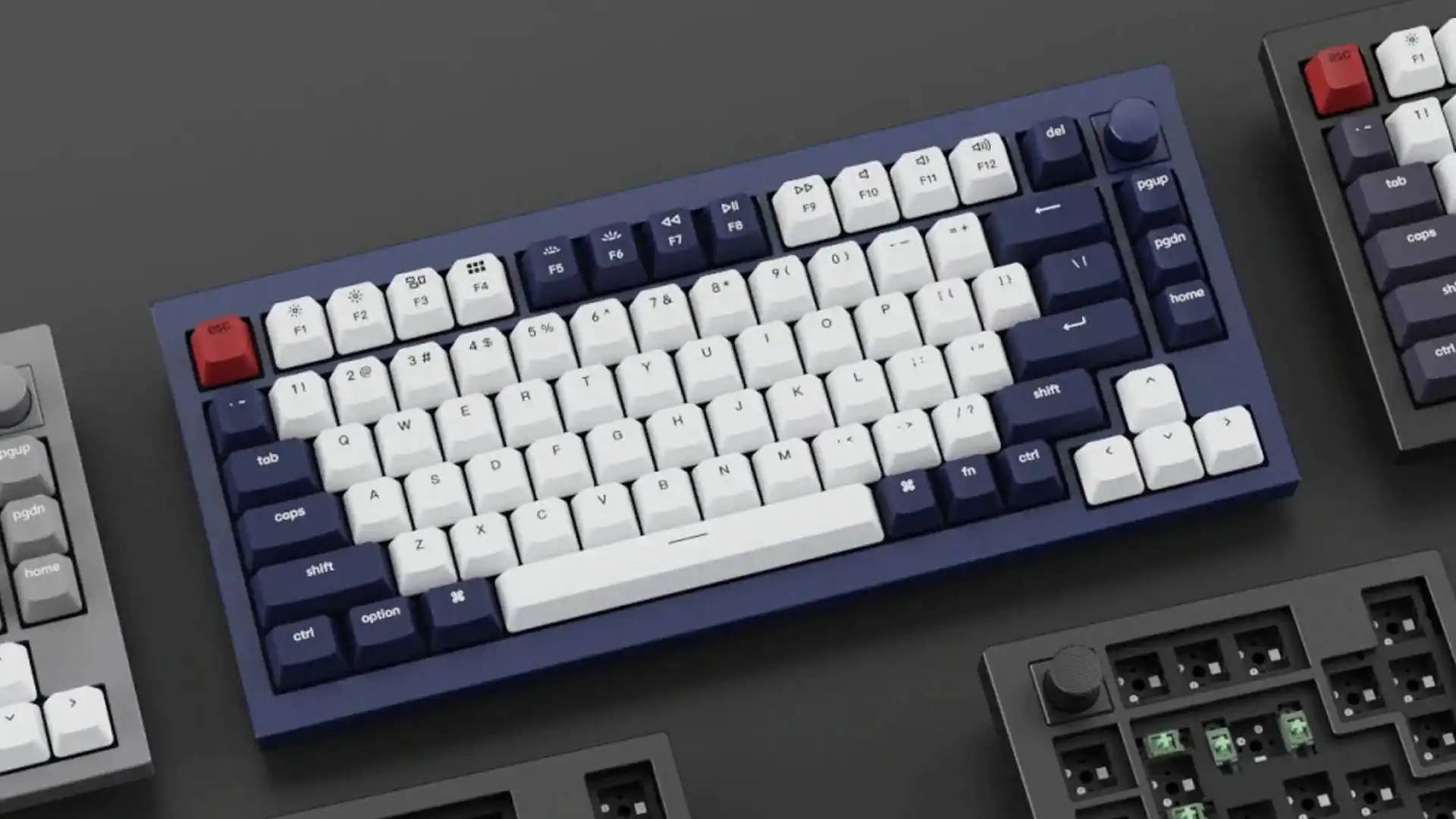 Navy blue and white compact mechanical keyboard with red escape key and rotary knob, surrounded by partial views of other tech accessories